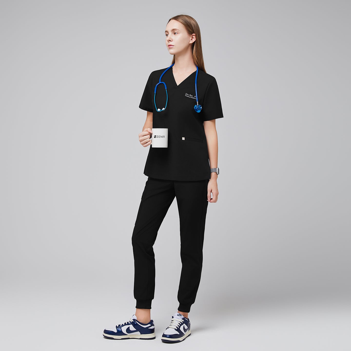 Image of Woman wearing a black V-neck scrub top with embroidered name and double front pockets, paired with matching black scrub pants,Eco Black